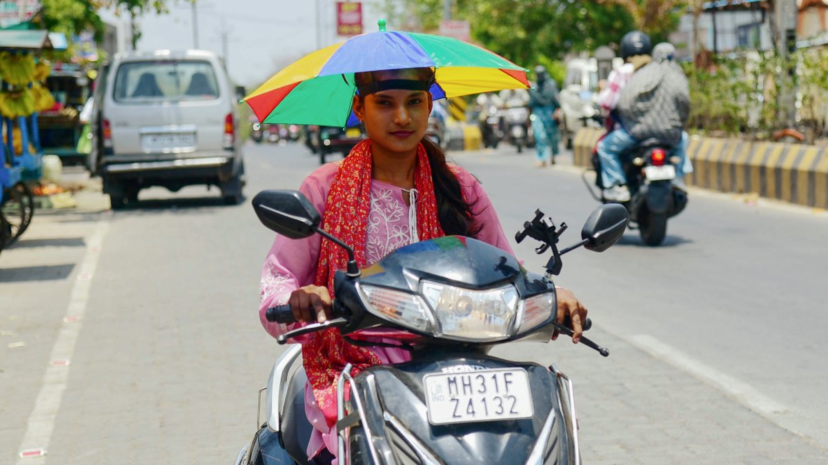 A woman rider on a scooty holds an umbrella over her head to protect herself…